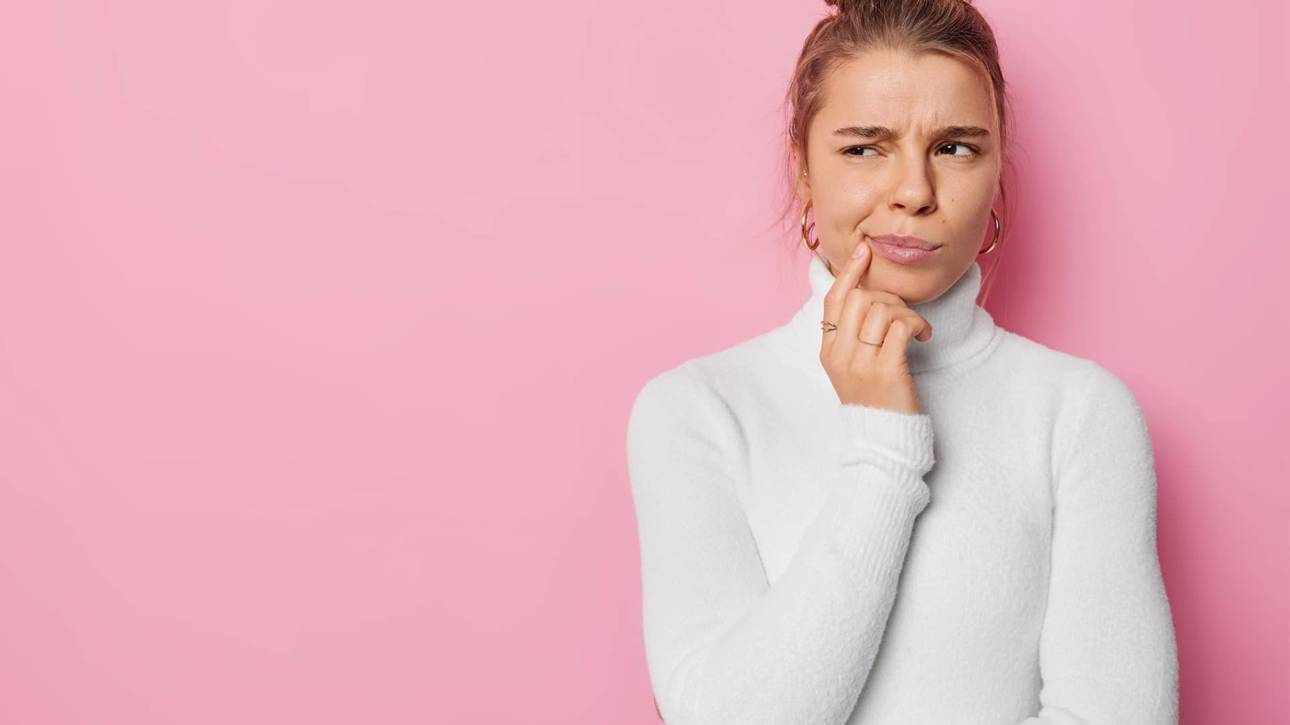 Ung kvinne i hvit genser ser tenksom ut og tar seg til kinnet, står foran rosa bakgrunn med skeptisk uttrykk. resorpsjon hos tannlegen tannen tannlegen roten behandling eksterne tid oslo hos universitetet lang